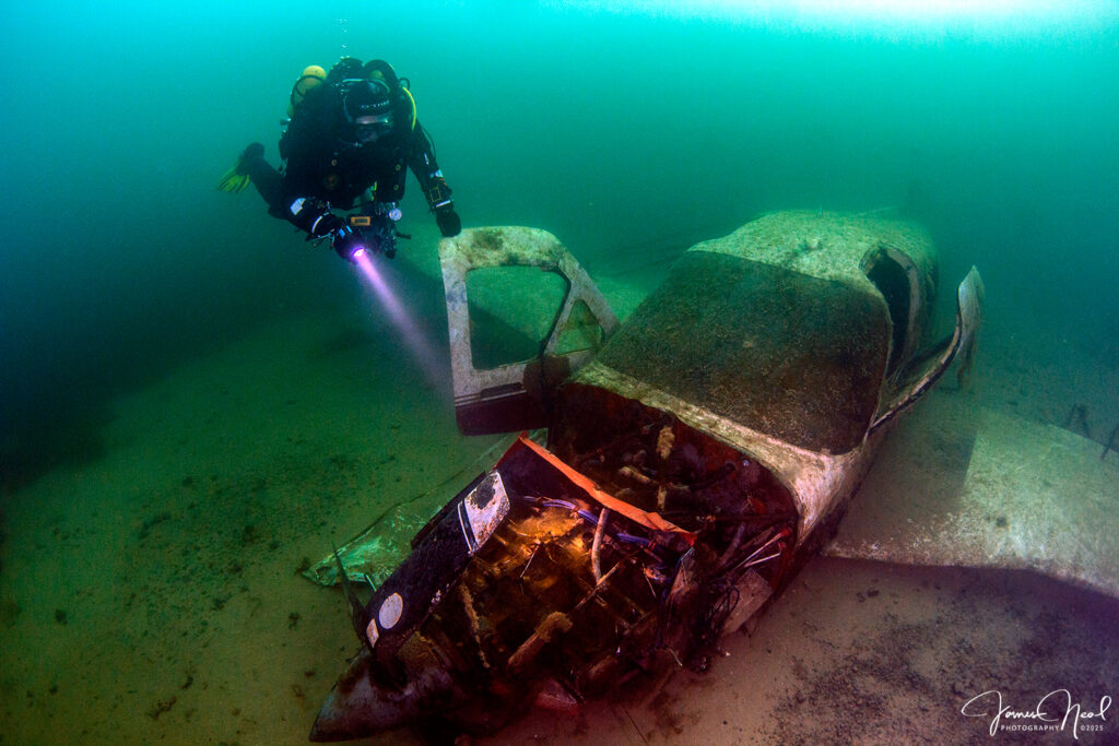 Underwater photograph of a scuba diver with a torch swimming towards the wreck of a light aircraft resting on the silty bottom of a quarry lake. The light of the torch casts a beam through the clear, greenish-blue water.