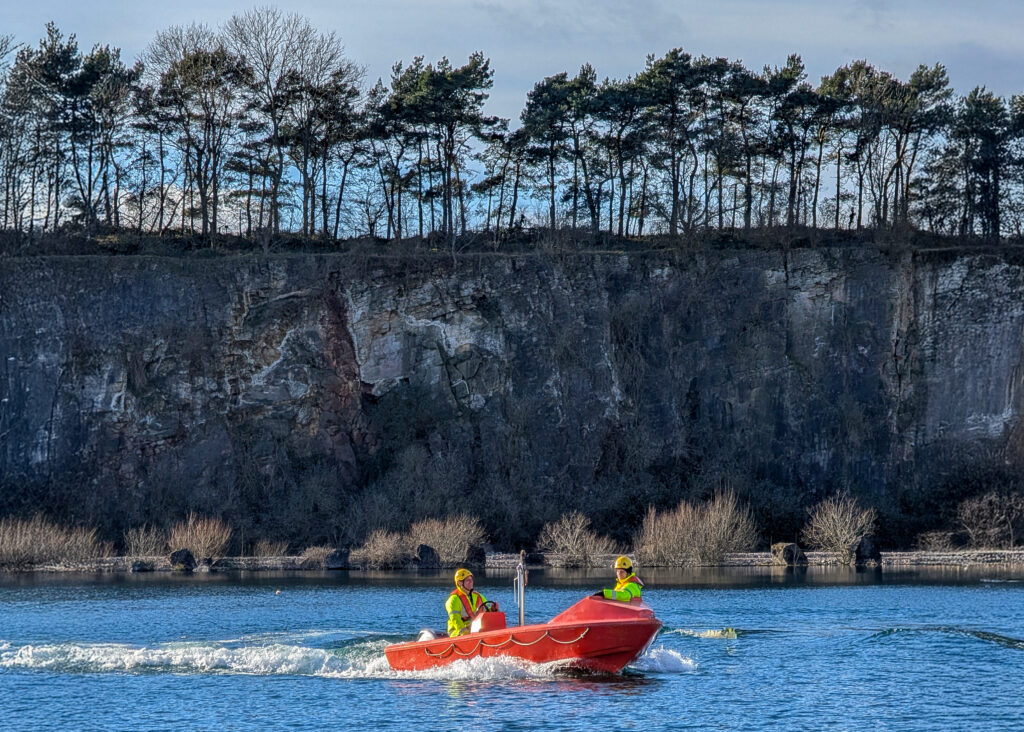 A rescue boat training exercise by South West Maritime Academy taking place in the water body at Cromhall Quarry. The cliff face in the background a mix of grey and tan colours, showing distinct layers, deep vertical cracks, and a rough, weathered texture.
