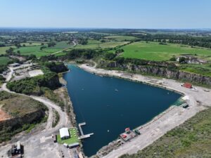 A wide, aerial photograph of Cromhall Quarry on a bright day. Sheer, layered rock cliffs surround a large, calm body of deep blue water. Green vegetation grows on the upper edges of the cliffs and fields surrounding the quarry.
